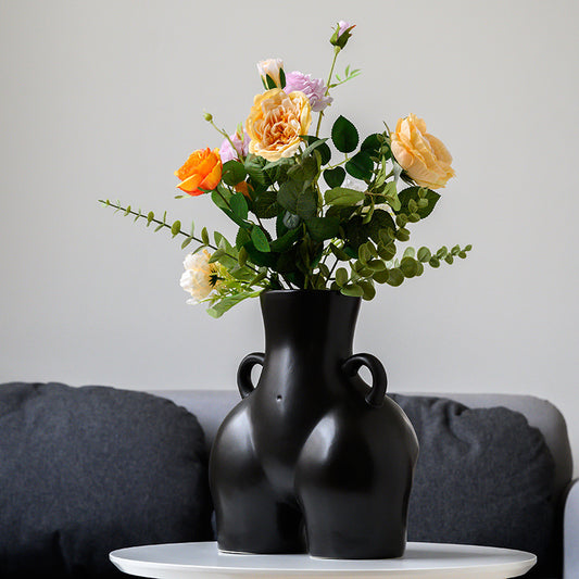 A black ceramic gourd-shaped vase on a white surface, with a bouquet of multicolored flowers arranged in it, placed against a neutral background with a grey sofa.