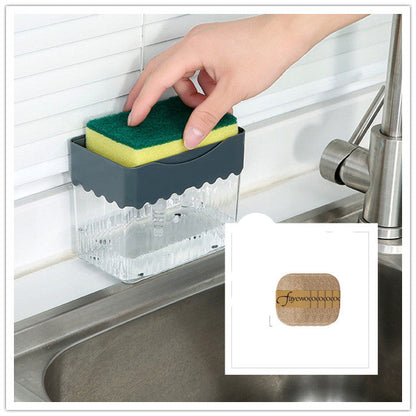 A person's hand pressing down on a sponge attached to a clear soap dispenser caddy, which is mounted on a wall beside a sink.