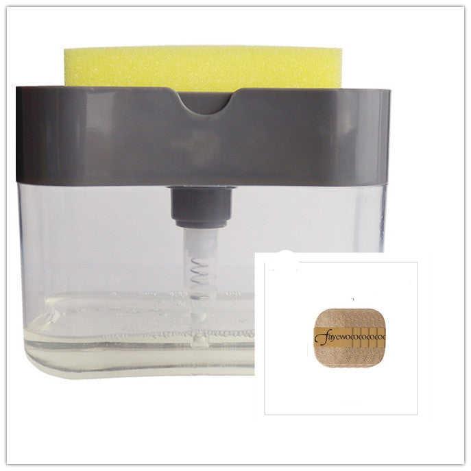 A person's hand pressing down on a sponge attached to a clear soap dispenser caddy, which is mounted on a wall beside a sink.