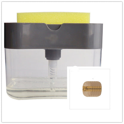 A person's hand pressing down on a sponge attached to a clear soap dispenser caddy, which is mounted on a wall beside a sink.