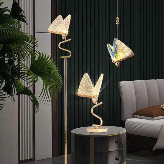 A modern floor lamp with a butterfly design, featuring gold-colored metal bodies and multicolored acrylic shades, standing in a room with a dark wall and a plant.