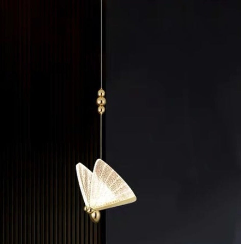 A modern floor lamp with a butterfly design, featuring gold-colored metal bodies and multicolored acrylic shades, standing in a room with a dark wall and a plant.
