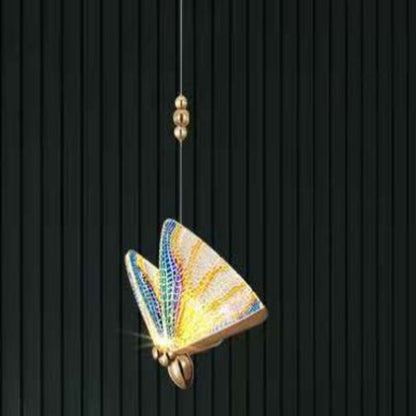 A modern floor lamp with a butterfly design, featuring gold-colored metal bodies and multicolored acrylic shades, standing in a room with a dark wall and a plant.