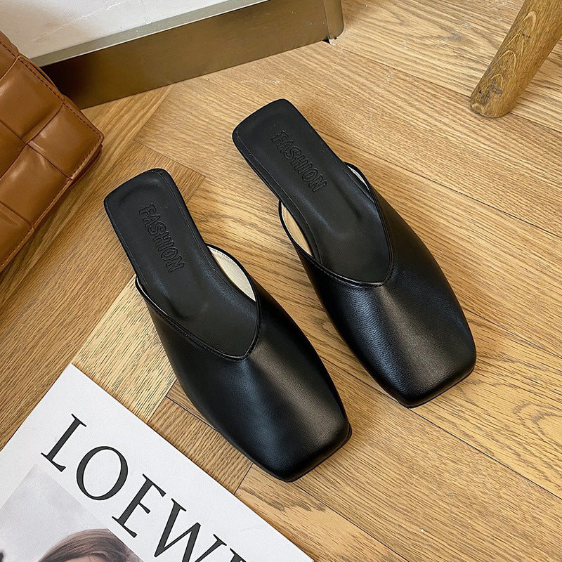 Black square-toe slippers on a wooden floor with a magazine underneath.