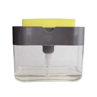 A person's hand pressing down on a sponge attached to a clear soap dispenser caddy, which is mounted on a wall beside a sink.