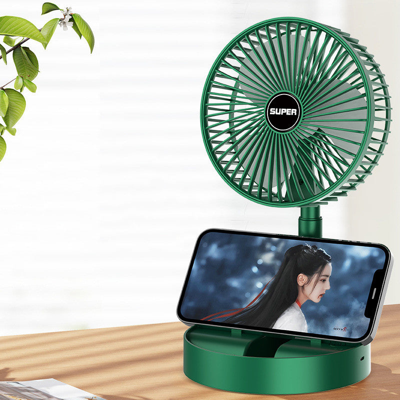 Green desk fan with smartphone holder on a wooden surface, plant in the corner.