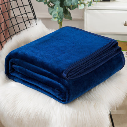 A blue coral fleece blanket placed on a white furry surface, with a part of a bed and a plant in the background.