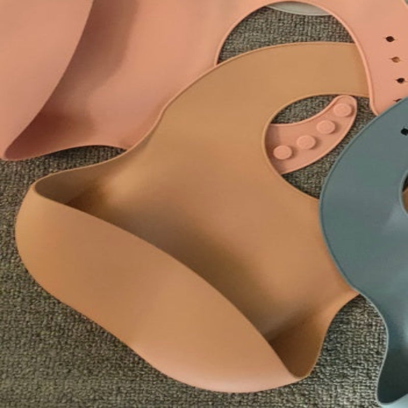 Three silicone bibs in pink, brown, and green on a textured surface