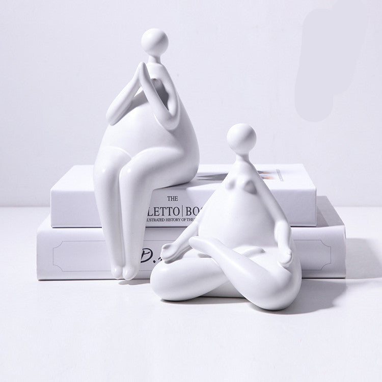 A pair of white ceramic figurines, one sitting and one standing, placed on top of a stack of books with a decorative vase beside them.