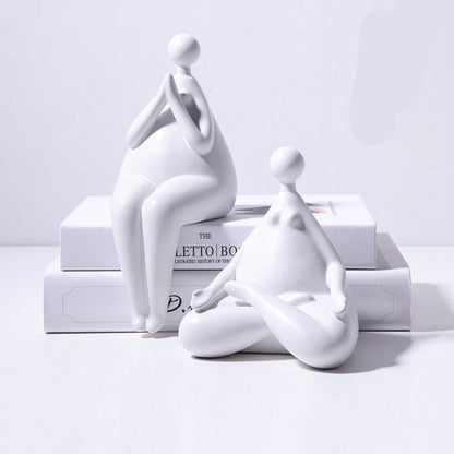 A pair of white ceramic figurines, one sitting and one standing, placed on top of a stack of books with a decorative vase beside them.
