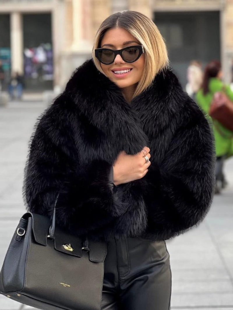 Woman wearing a black fur coat and sunglasses, holding a black handbag.