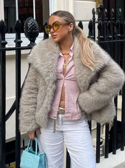 Woman wearing a gray fur coat, pink shirt, and white pants standing in front of a black iron gate.