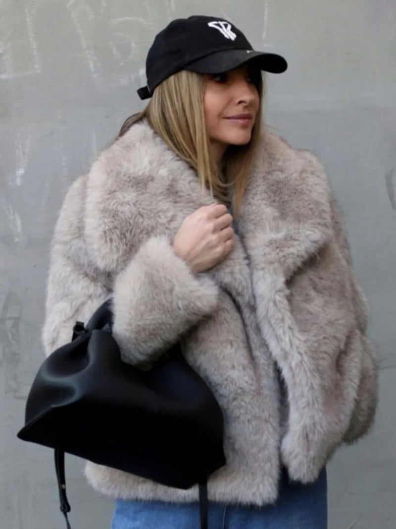 Person wearing a fluffy gray coat and black cap, holding a black bag against a gray background