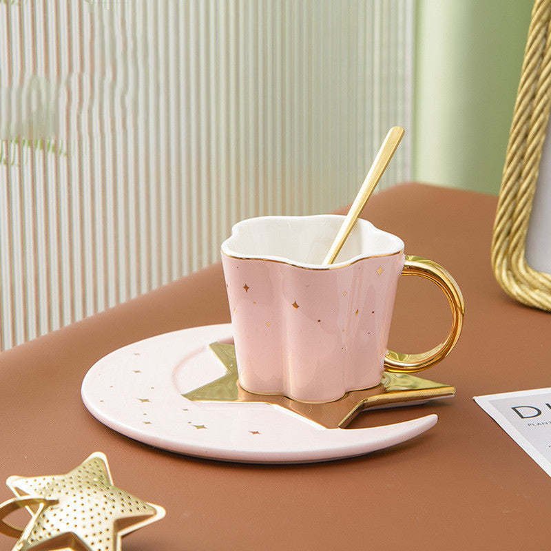 A set of ceramic cups with star and moon saucers in black, white, and gold colors, displayed on a table with books and decorative stars.
