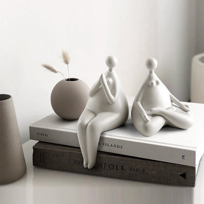 A pair of white ceramic figurines, one sitting and one standing, placed on top of a stack of books with a decorative vase beside them.