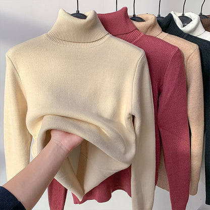 Beige turtleneck sweater held by a hand with other sweaters in the background.