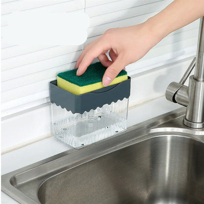 A person's hand pressing down on a sponge attached to a clear soap dispenser caddy, which is mounted on a wall beside a sink.