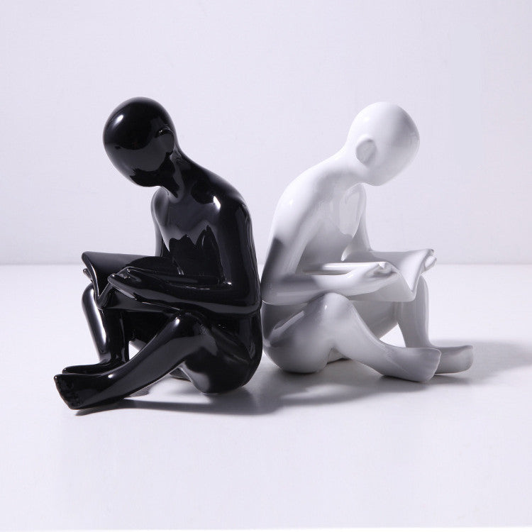 A pair of white ceramic figurines, one sitting and one standing, placed on top of a stack of books with a decorative vase beside them.