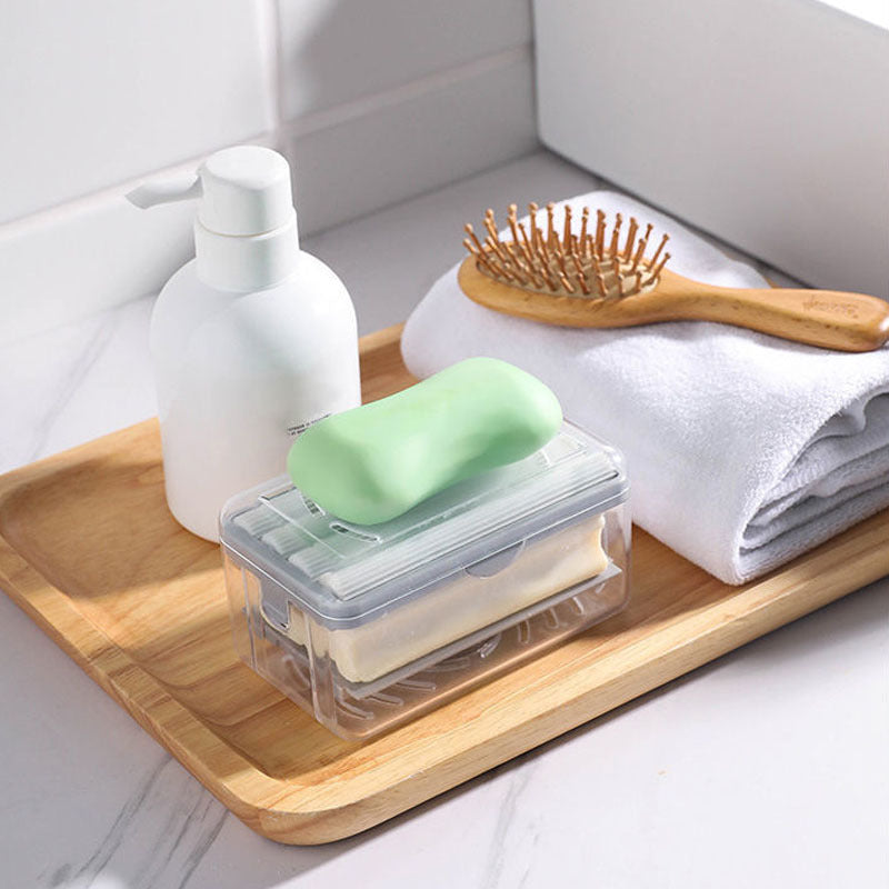 Clear soap container with a bar of soap on a wooden tray with a bottle, brush, and towel in the background.