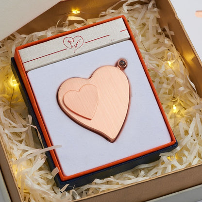 Heart-shaped copper pendant in a decorative box with white shreds and string lights in the background