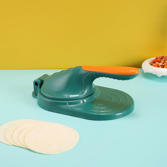 Green tortilla press with orange handle on a blue surface with tortillas and a bowl of ingredients.